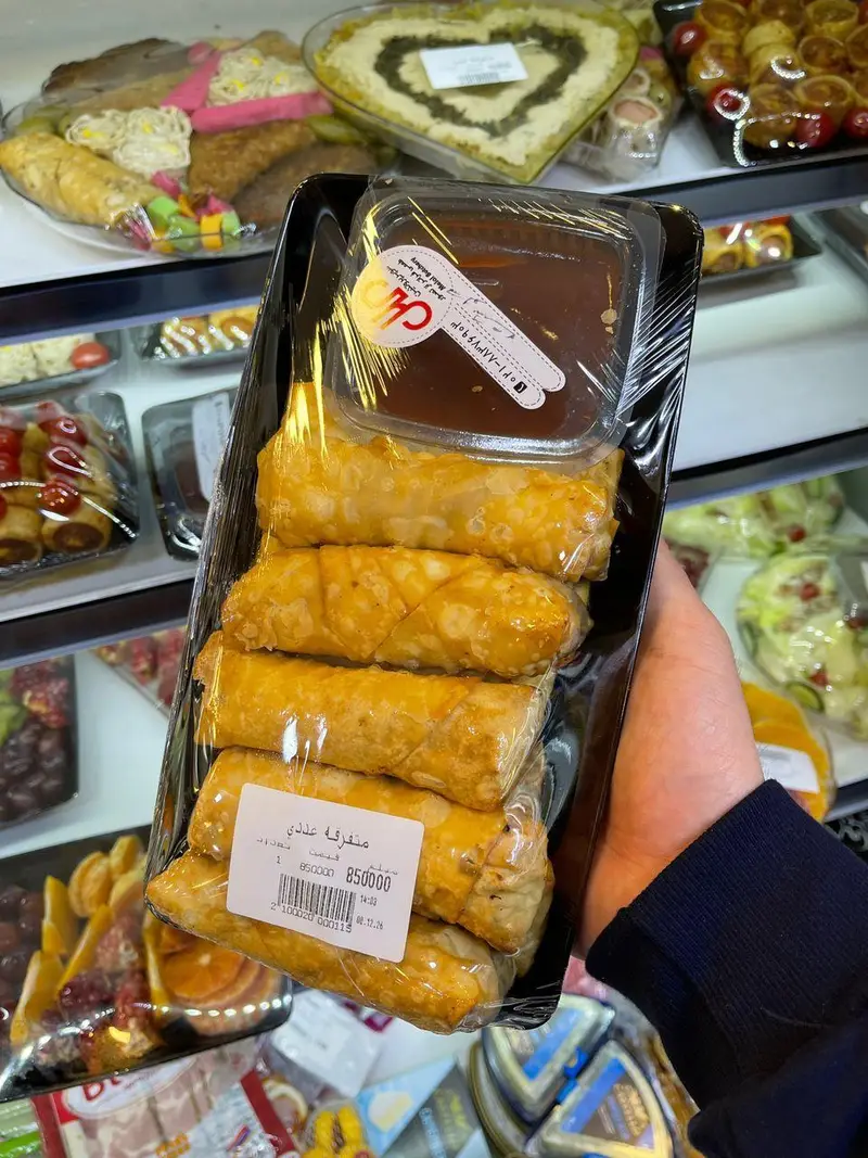 بسته بندی شیرینی رول طلایی با سس، در دست مشتری داخل قفسه‌های متنوع فروشگاه سوپر پروتئین ملل. محصولی آماده با قیمت درج شده.