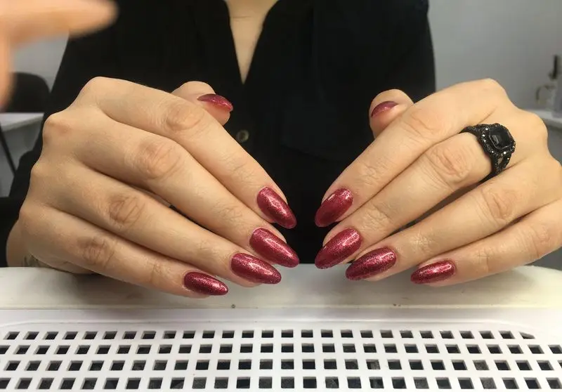 طراحی و کاشت ناخن با لاک اکلیلی قرمز تیره و درخشان روی دست‌های یک زن همراه با انگشتر مشکی در مرکز زیبایی مریم موسوی