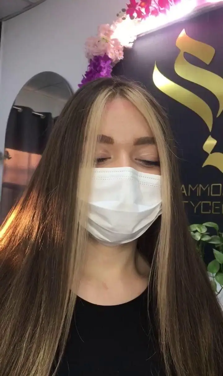 مشتری با ماسک در مریم موسوی بیوتی سنتر با موهای بلند قهوه ای و فیس فریم بلوند، نشان دهنده خدمات رنگ و لایت مو در سالن.