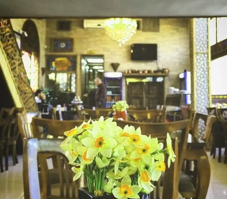 فضای داخلی دلنشین رستوران خوان باشی با میز و صندلی چوبی، لوستر زیبا و گلدان گل نرگس که فضایی گرم و سنتی را به نمایش می گذارد.