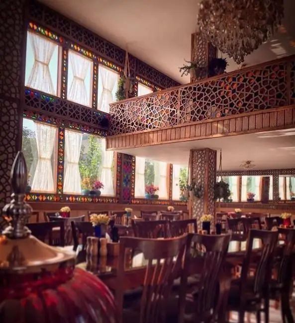 نمای داخلی رستوران خوان باشی با دکوراسیون سنتی و معماری زیبای ایرانی، شامل پنجره‌های رنگی و میز و صندلی‌های چوبی، فضایی دلنشین برای غذاخوری.