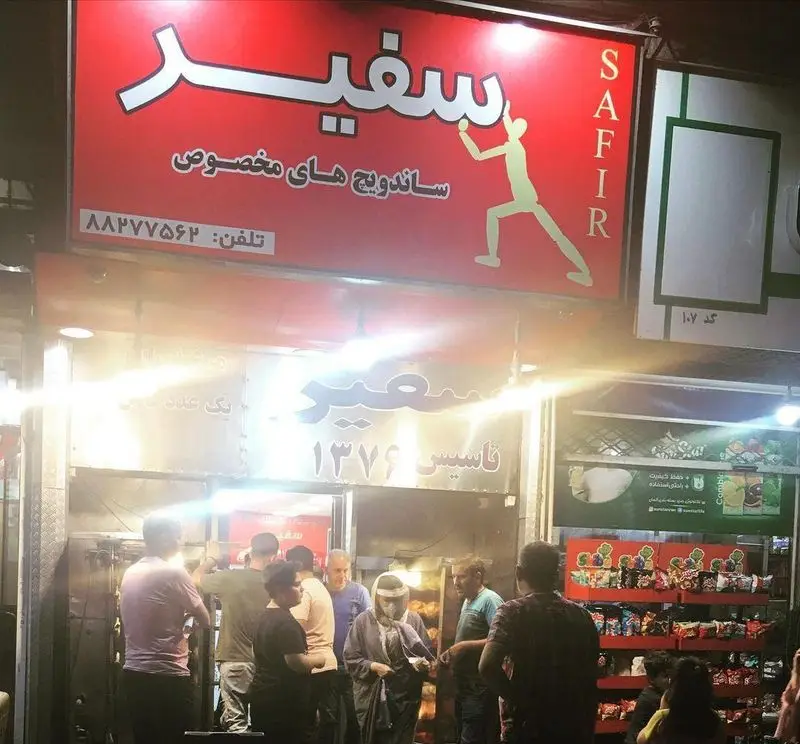تصویر فست فود سفیر، با تابلوی قرمز رنگ و لوگوی مرد در حال پرش، ارائه‌دهنده ساندویچ‌های مخصوص. تأسیس ۱۳۷۶ و تلفن ۸۸۷۷۷۵۶۲.