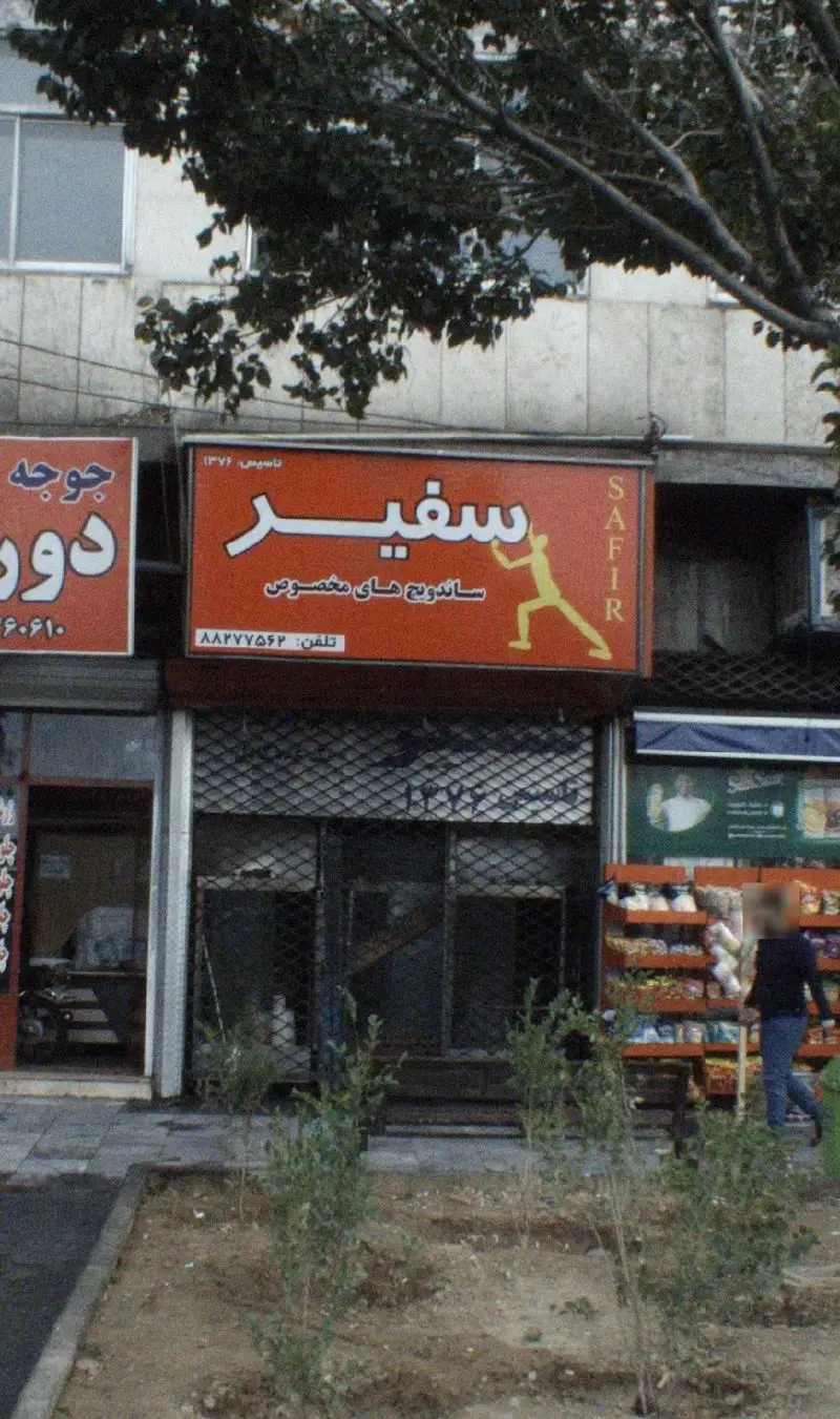 تابلوی قرمز رنگ فست فود سفیر با حروف فارسی و لاتین SAFIR، تبلیغ ساندویچ های مخصوص و لوگوی ورزشکار در نمای بیرونی.