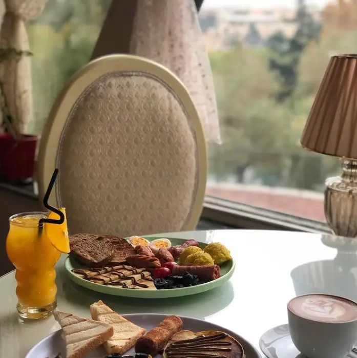 صبحانه کامل و لذیذ کافه سینان شامل پلیت‌های متنوع نان، سوسیس، تخم مرغ، زیتون، قهوه و آب پرتقال با منظره‌ای دلنشین.