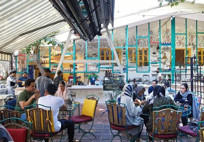 محیط دنج و رنگارنگ کافه سینان با مشتریانی که در فضای باز زیر سایه بان از نوشیدنی و گفتگو لذت میبرند، دکوراسیون خاص با پنجره‌های آبی و زرد.