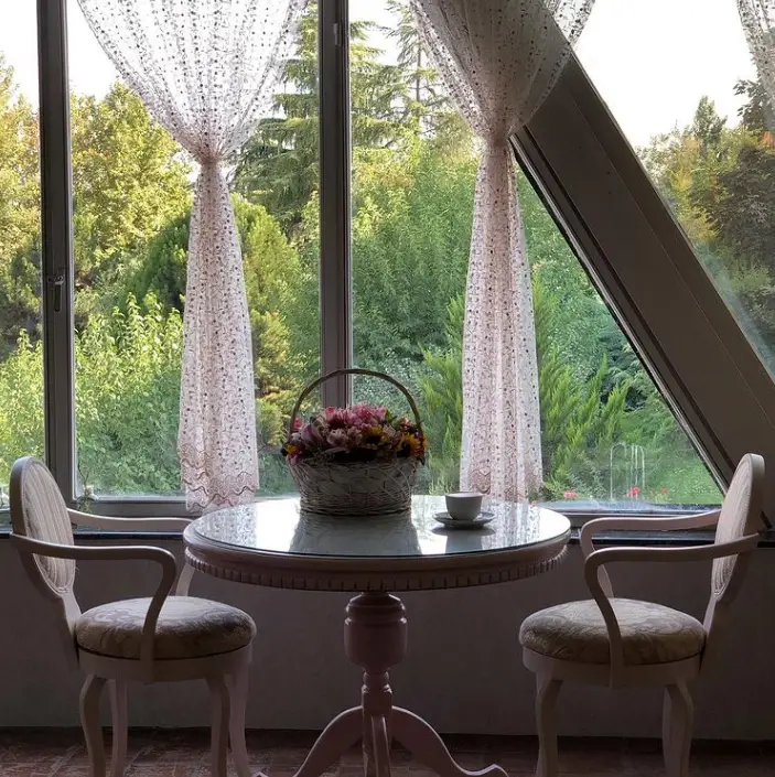 کافه سینان: نمایی دلنشین از میز شیشه‌ای، سبد گل، فنجان چای و صندلی‌ها کنار پنجره بزرگ با چشم‌انداز درختان سرسبز.