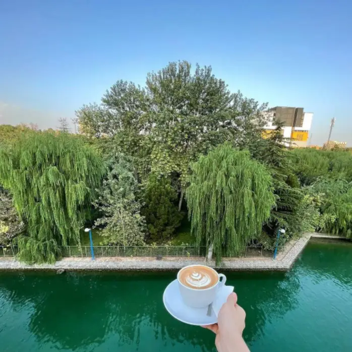 نمایی زیبا از یک فنجان لاته خوش‌طعم با طرح هنری بر روی آن، در دست مشتری کافه سینان، مشرف به منظره‌ای دلنشین از طبیعت سرسبز و آب آرام.