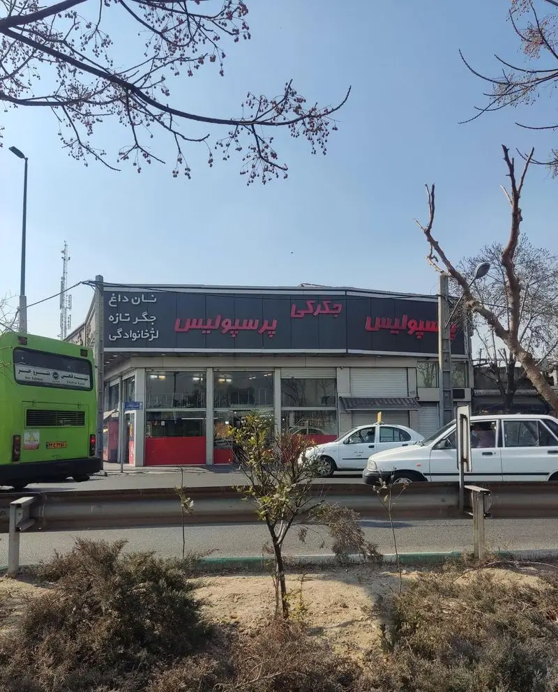 نمای خارجی جگرکی پرسپولیس با تابلو قرمز و مشکی، همراه با اتوبوس و خودروها در خیابان. ارائه جگر تازه و نان داغ.