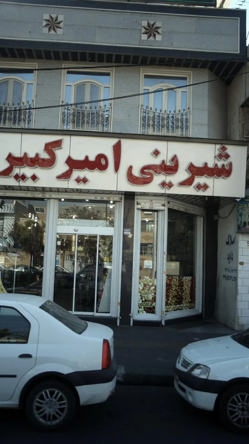نمای خارجی شیرینی فروشی امیرکبیر با تابلوی قرمز، پنجره‌های ویترین حاوی شیرینی و آجیل و ماشین‌های پارک شده.