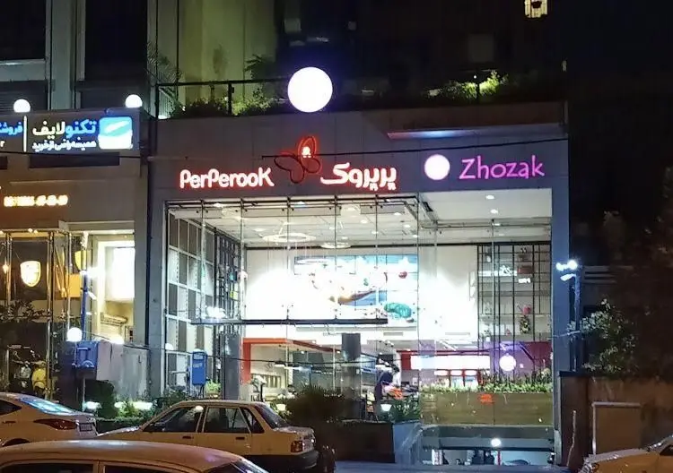 نمای شبانه و جذاب فست فود پرپروک با تابلوی روشن و نماد پروانه، ورودی شیشه‌ای مدرن، در یک خیابان شلوغ با ماشین‌ها.