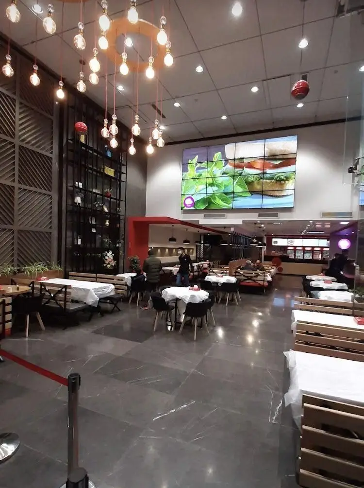 فضای داخلی مدرن و شیک رستوران پرپروک با میز و صندلی‌های چوبی، لوستر زیبا و نمایشگر بزرگ غذای ساندویچ و سالاد.