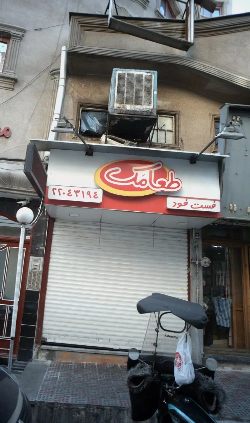 نمایی از فست فود طعامک با تابلوی قرمز و زرد جذاب، شامل شماره تماس ٢٢.٤٣١٩٤، کرکره بسته و یک موتورسیکلت در جلوی مغازه.