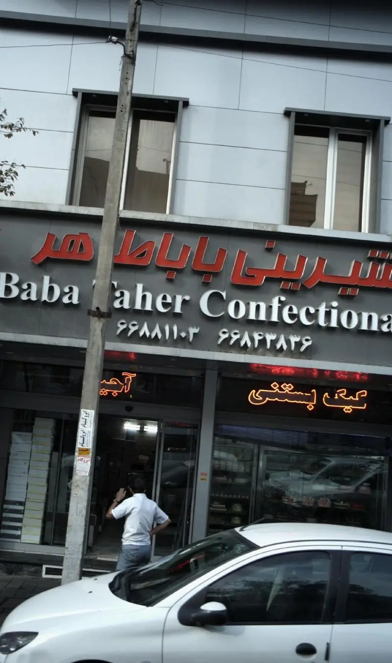 تصویر نمای بیرونی شیرینی‌فروشی باباطاهر با تابلوهای فارسی و انگلیسی، شماره‌های تماس، و ورودی مغازه همراه با یک خودروی سفید