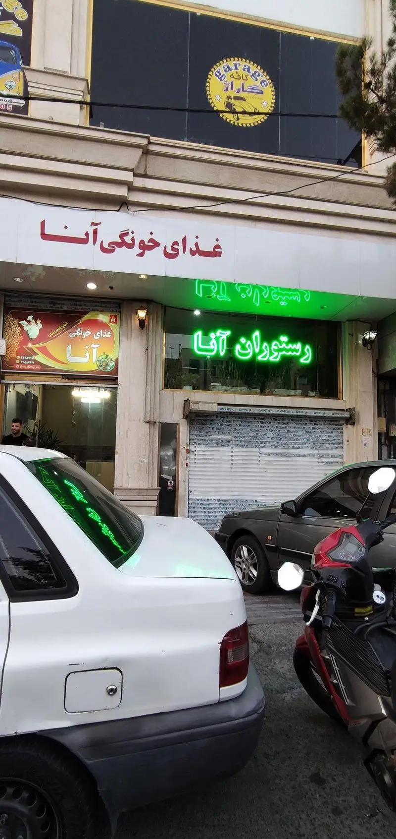 نمای بیرونی کسب و کار غذا خونگی آنا، رستوران آنا با تابلوهای نئون سبز و بنر تبلیغاتی، کافه گاراژ در بالا.