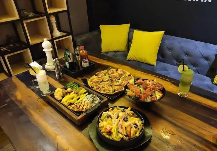 میز پر از غذاهای کافه رستوران پرشین: پیتزا، بشقاب استیک و سیب‌زمینی، سالاد و پاستا با نوشیدنی در محیطی شیک.