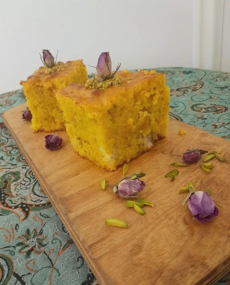 دو برش کیک زعفرانی زرد و خوشمزه، تزئین شده با غنچه گل محمدی بنفش و پسته سبز روی تخته چوبی. دسر خاص گروه تشریفاتی لیمون برای مراسم شما.
