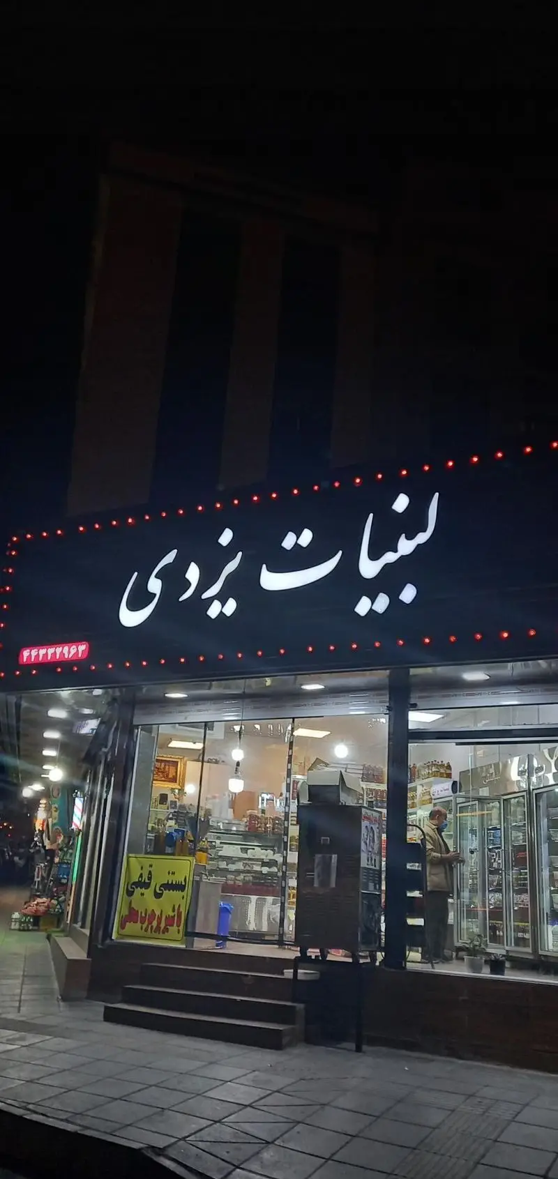 عکس شبانه از مغازه لبنیات یزدی شهران با تابلوی نئون درخشان، ارائه دهنده انواع محصولات لبنی سنتی و بستنی قیفی، محیطی دلنشین.