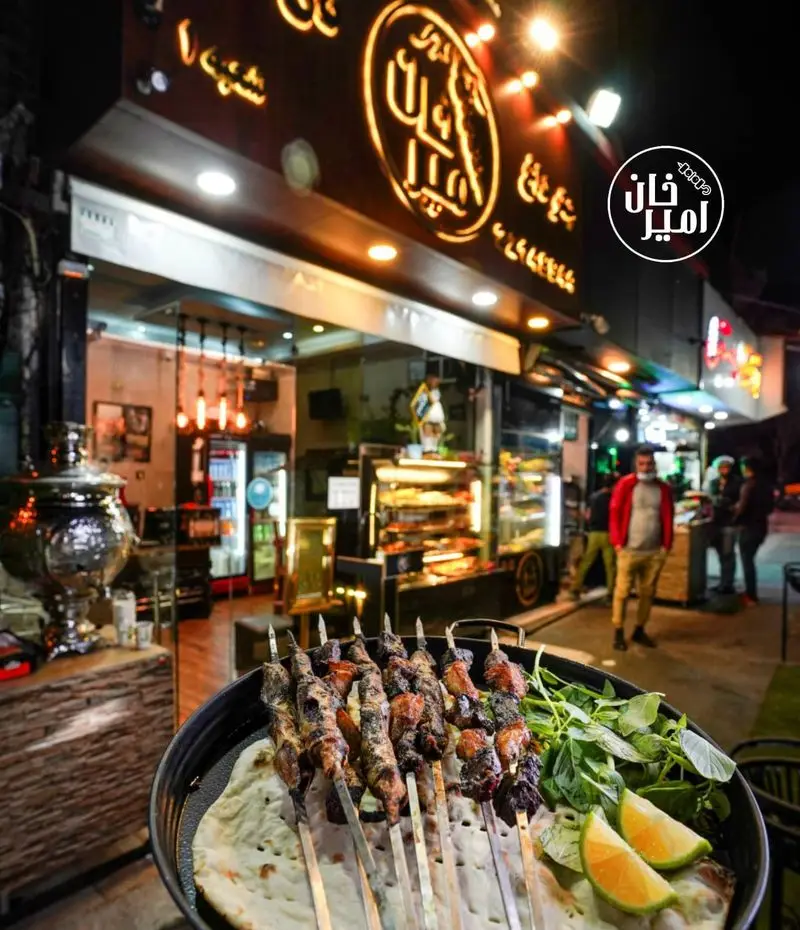 عکس از نمای شب جگرسرای امیرخان (گل یخ) و سیخ‌های لذیذ کباب جگر و دل و قلوه روی نان و سبزی تازه.