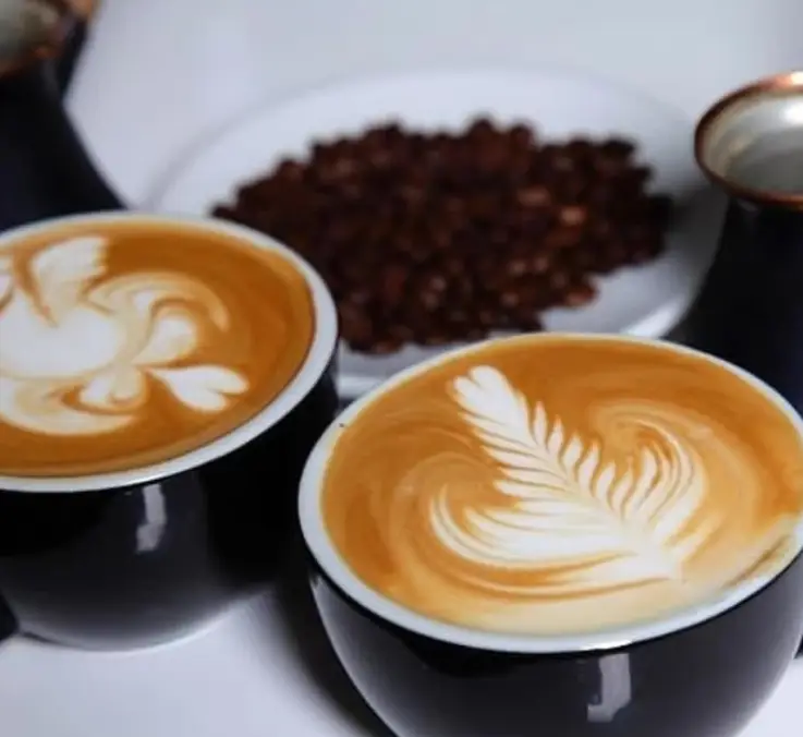 دو فنجان قهوه لاته آرت حرفه‌ای با طرح‌های دلنشین قو و برگ در کافه تی وانا، کنار دانه‌های تازه قهوه. بهترین نوشیدنی گرم برای شما.