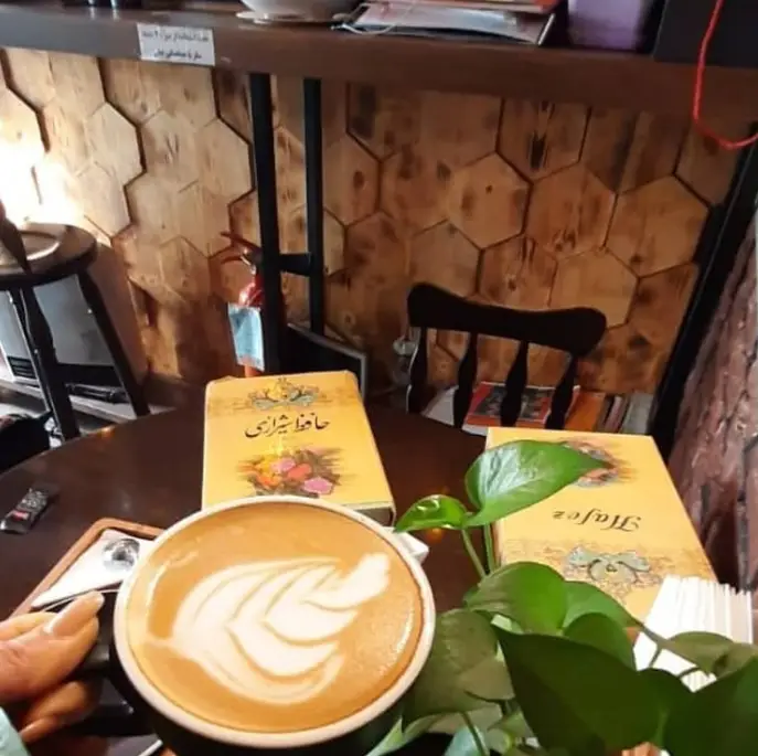 لذت نوشیدن لاته با طرح هنری در کافه تی وانا، میز چوبی با دیوان حافظ شیرازی و گلدان سبز. محیطی دلنشین برای مطالعه و آرامش.