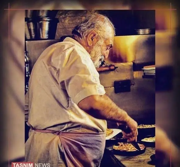 پیرمرد آشپز باتجربه و مهربان کافه سامیا، با ریش سفید و پیراهن سفید، در حال پخت و پز غذاهای لذیذ در آشپزخانه گرم و صمیمی