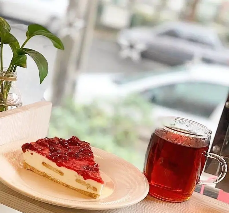 تصویری جذاب از یک برش چیزکیک با رویه توت‌فرنگی یا گیلاس و چای گرم در کافه سامیا، ایده‌آل برای یک عصر دلنشین با منظره شهری.