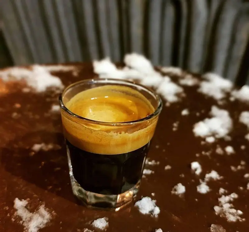 شات اسپرسوی کافه سامیا: نوشیدنی گرم و دلپذیر با کرمای غنی و طلایی، روی میز چوبی مزین به دانه‌های سفید، آماده برای شما. لذت قهوه با کیفیت.