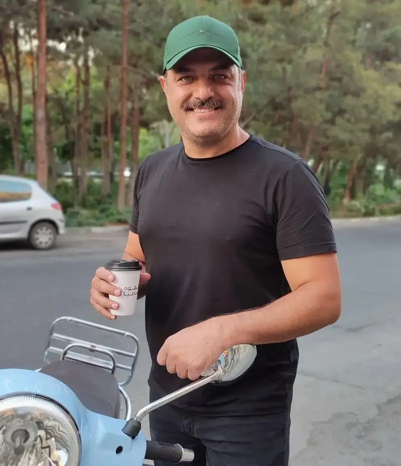مردی شاد با کلاه سبز و تی‌شرت مشکی، قهوه بیرون بر کافه سامیا را در دست گرفته و کنار اسکوتر آبی رنگ خود لبخند می‌زند.