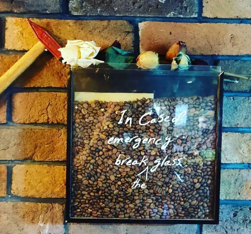 تصویر جذاب از جعبه شوخی "قهوه اضطراری" پر از دانه های قهوه در کافه سامیا، با تابلو "در مواقع اضطراری شیشه را بشکنید" و تبر کوچک.