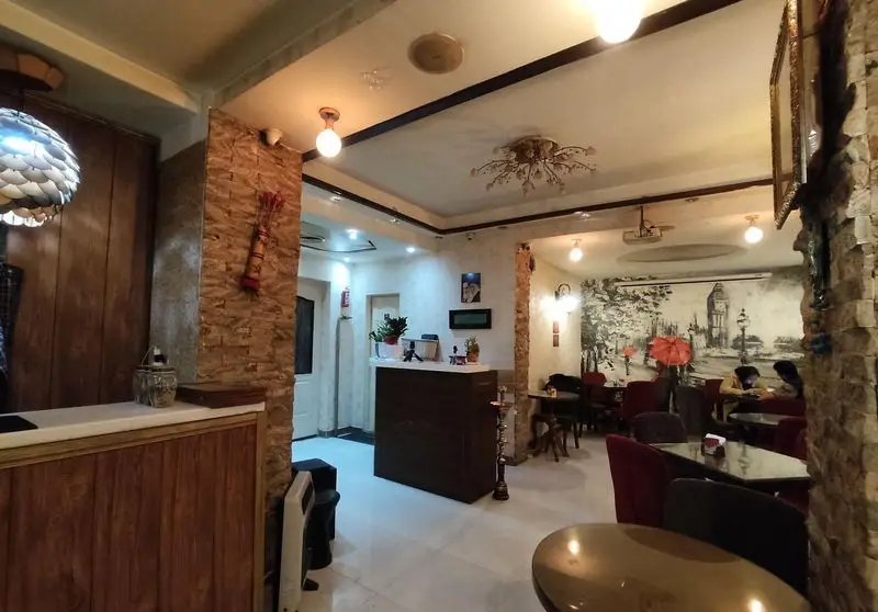 نمایی دلنشین از فضای داخلی کافه شاپرک: دکوراسیون سنگی، میز پذیرش چوبی، نورپردازی گرم و نقاشی دیواری زیبای لندن روی دیوار اصلی.