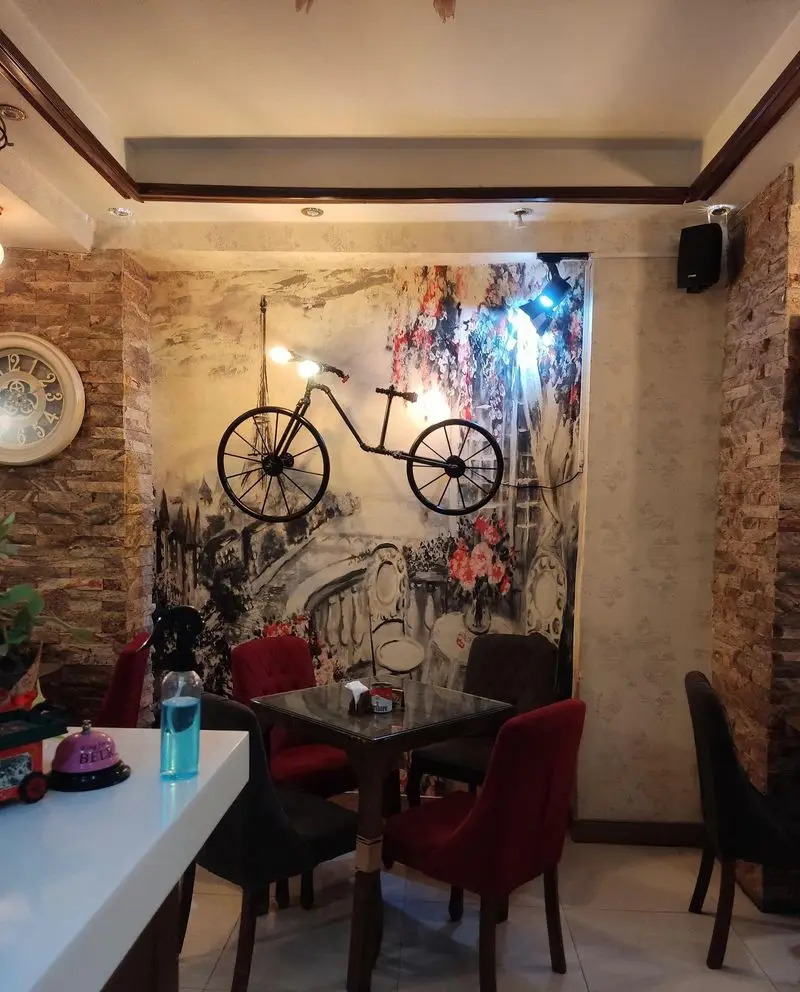 فضای داخلی کافه شاپرک با دکوراسیون خاص شامل دیوار آجری، نقاشی دیواری با دوچرخه نورانی، میزهای قهوه‌ای و صندلی‌های قرمز و خاکستری.