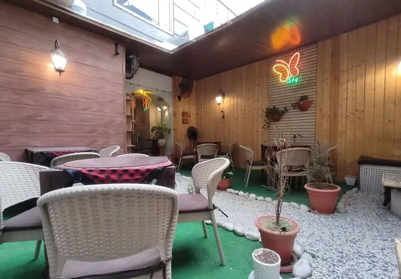 فضای حیاط دنج کافه شاپرک با دکوراسیون چوبی، میز و صندلی‌های حصیری، تابلوی نئون شاپرک و مسیر سنگی.