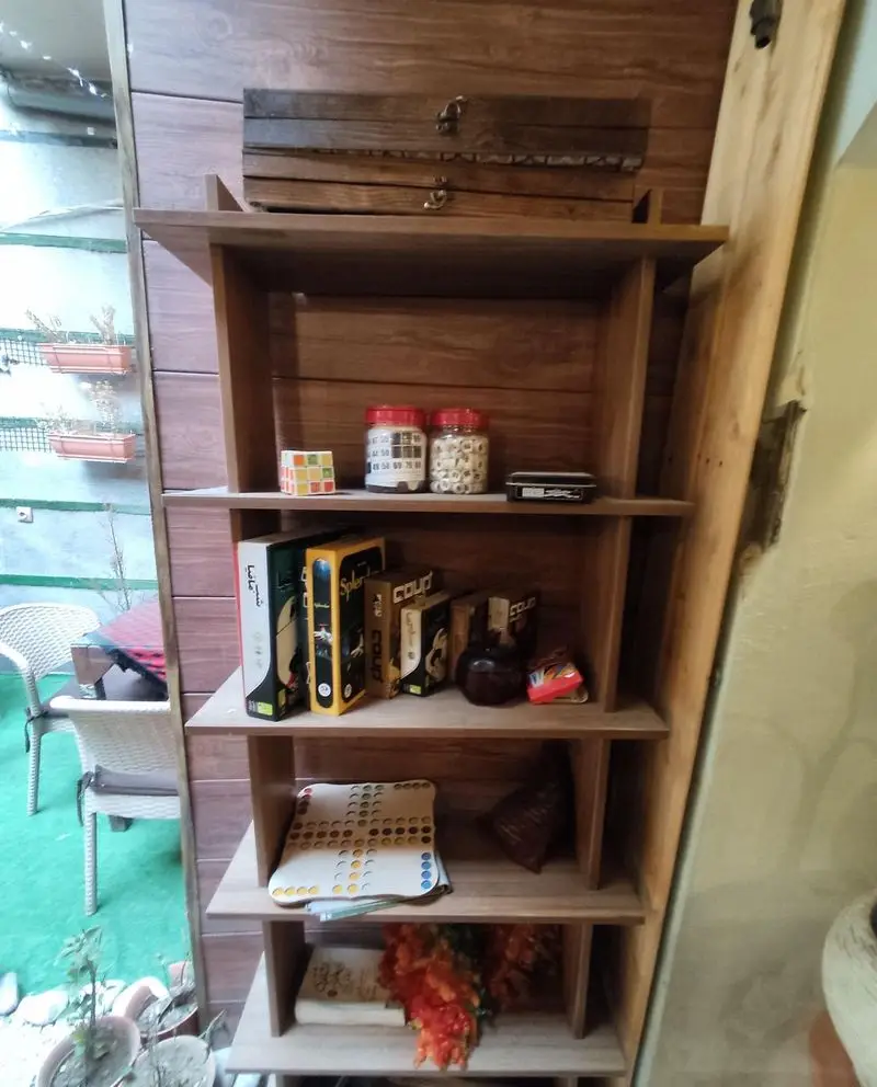 قفسه چوبی زیبای کافه شاپرک با مجموعه‌ای از بازی‌های رومیزی جذاب، کتاب و دکوری‌های خاص، ایجاد فضایی دنج