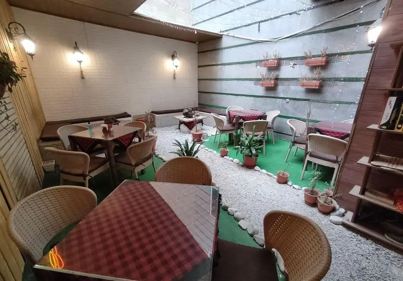 فضای دلنشین و سرسبز کافه شاپرک با میز و صندلی‌های حصیری، مسیر سنگی، و گلدان‌های تزئینی، مناسب برای دورهمی دوستانه.