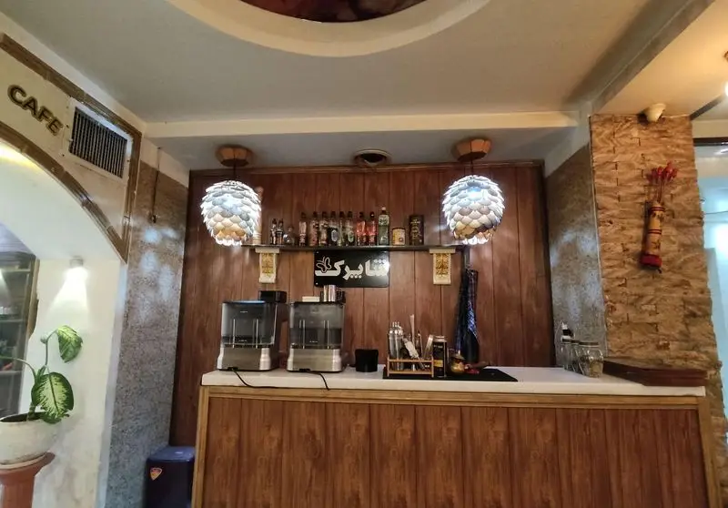 دکوراسیون داخلی و فضای گرم و دلنشین کافه شاپرک، شامل پیشخوان بار مجهز به دستگاه‌های نوشیدنی و لامپ‌های تزئینی زیبا.