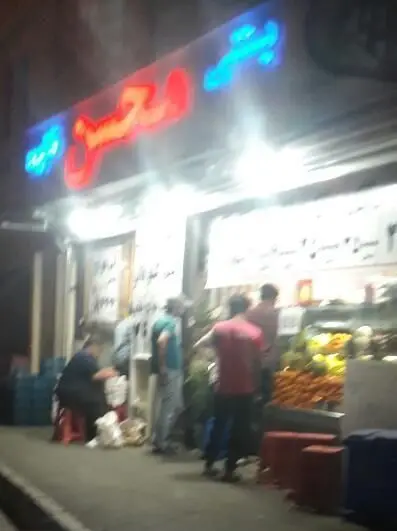 تصویری از بستنی محسن در شب با تابلو نئون روشن، مشتریان در حال خرید آبمیوه و بستنی تازه. میوه‌های رنگارنگ در ویترین.