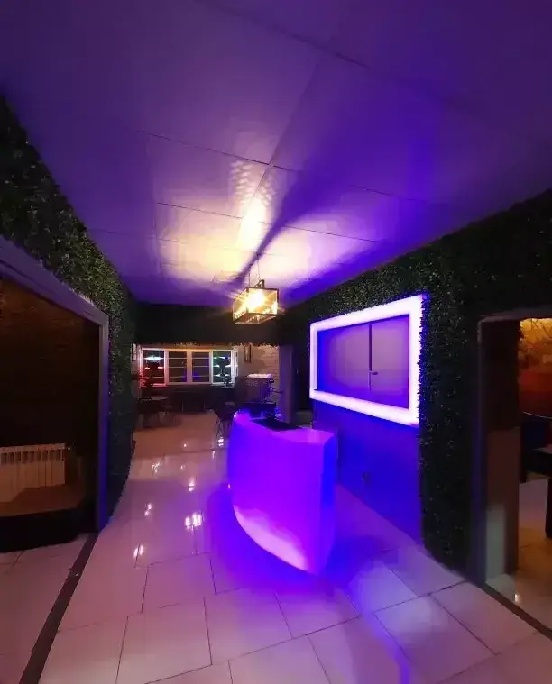 ورودی مدرن کافه قلیان عربی A&M با نورپردازی بنفش خاص، کانتر پذیرش درخشان و دیوارهای سبز پوشیده از گیاه.