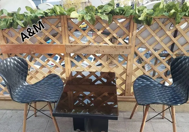 فضای باز دلنشین کافه قلیان عربی A&M: میز و صندلی مدرن مشکی، پارتیشن چوبی زیبا با تزئین برگ‌های سبز مصنوعی برای مشتریان.