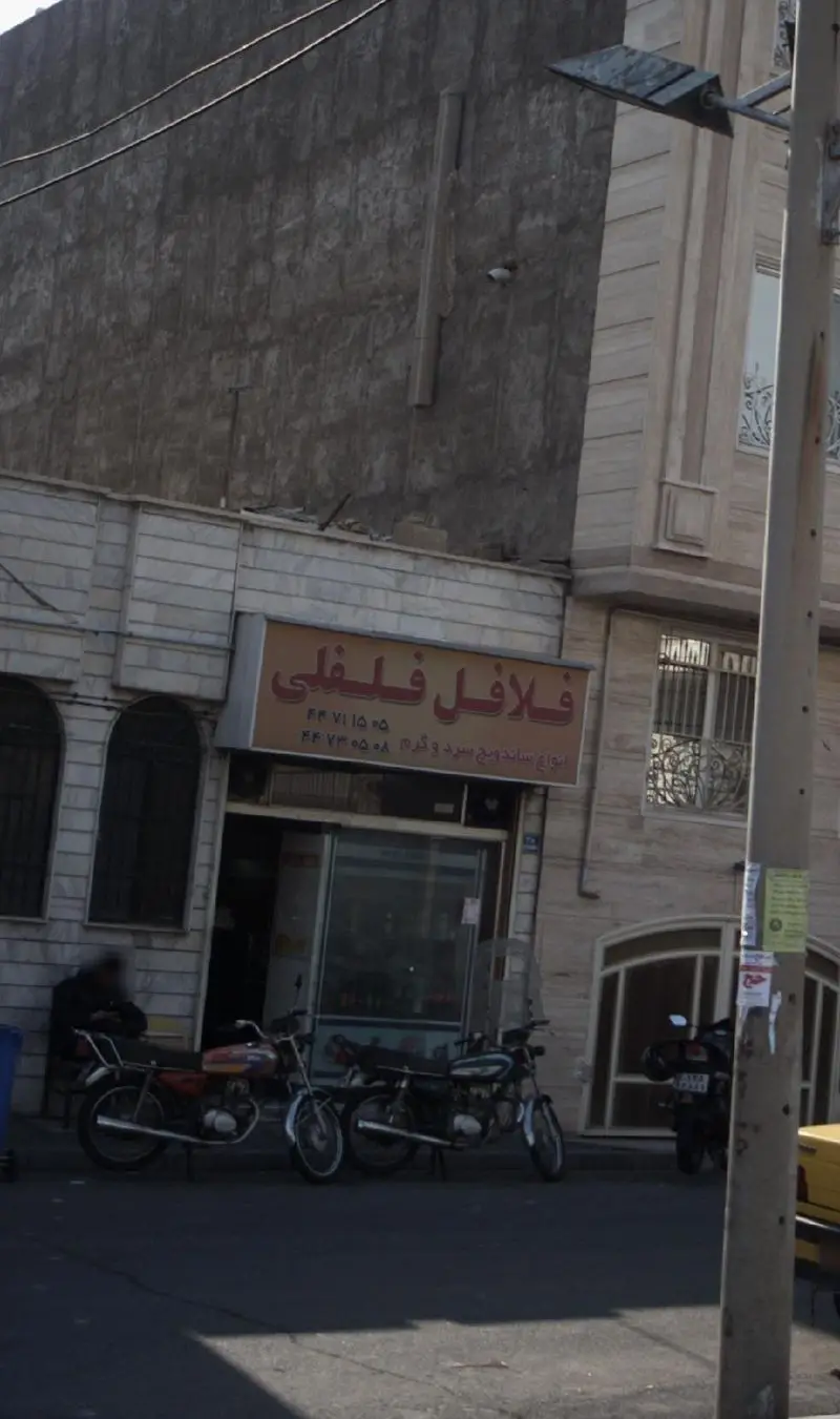 تصویر نمای بیرونی مغازه غذایی فلافل فلفلی در خیابان، با تابلو نارنجی رنگ ساندویچ سرد و گرم، شماره تلفن و موتورسیکلت‌های مشتریان.