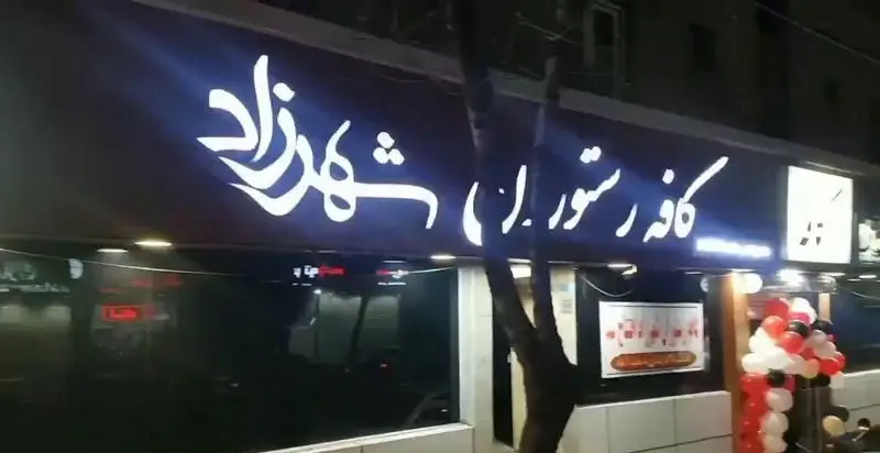 نمای بیرونی کافه رستوران شهرزاد در شب با تابلو نورانی و دکوراسیون ورودی شامل بادکنک‌های رنگی، فضایی دلنشین و دعوت‌کننده برای مشتریان.