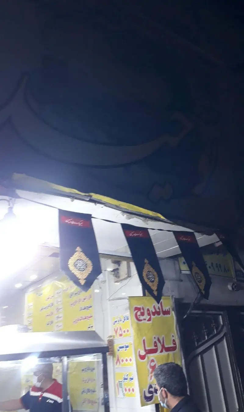 نمایی شبانه و دلنشین از پیتزا ساندویچ توحید، مزین به پرچم‌های مذهبی. تابلوی روشن ساندویچ فلافل و فضای گرم خدمت‌رسانی به مشتریان.