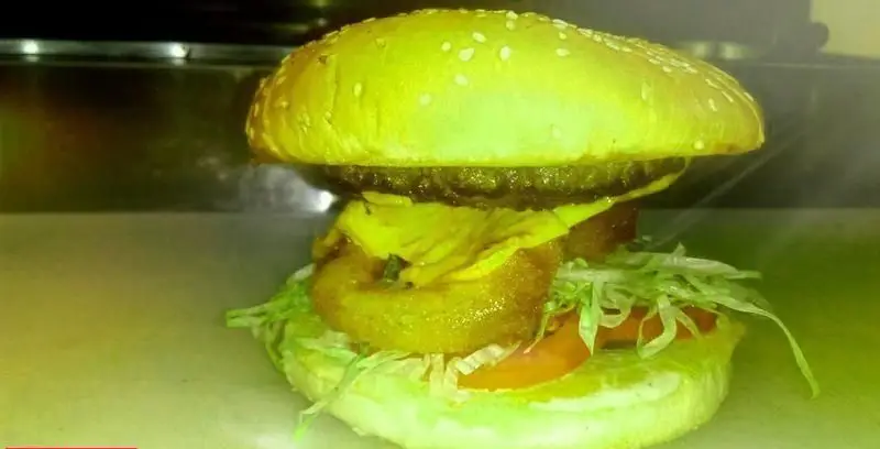 تصویری جذاب از یک همبرگر خوشمزه و کامل از فست فود شورینه، شامل نان کنجدی، پتی گوشت، پنیر آب شده، پیاز سوخاری، کاهو و گوجه.