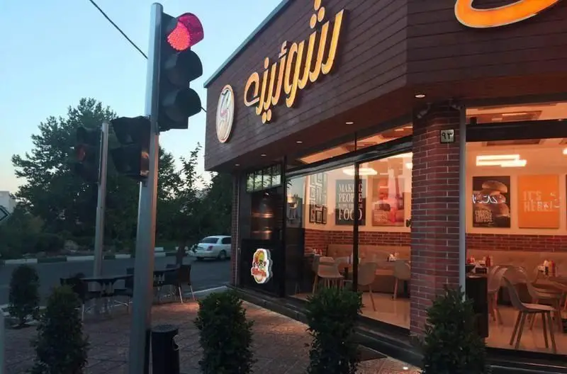 تصویر نمای بیرونی فست فود شورینه در یک غروب دل‌انگیز، با تابلوی نورانی، چراغ راهنمایی قرمز و فضای داخلی مدرن و میزهای چیده‌شده.