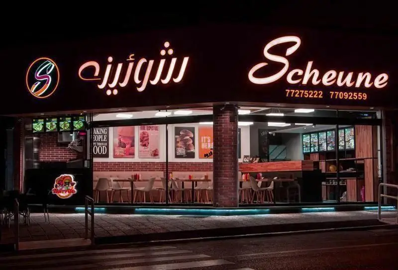 نمای شب از فست فود شورینه (Scheune) با تابلوی نئون روشن و لوگو S. داخل رستوران با میز و صندلی و پوسترهای غذایی جذاب. شماره تماس: 77275222.
