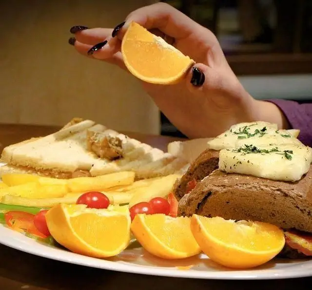 نمایی از دست مشتری که پرتقال را روی بشقاب صبحانه یا ناهار کامل کافه داریما می‌فشارد. شامل ساندویچ، سیب‌زمینی و نان همراه با پنیر و سبزیجات تازه.