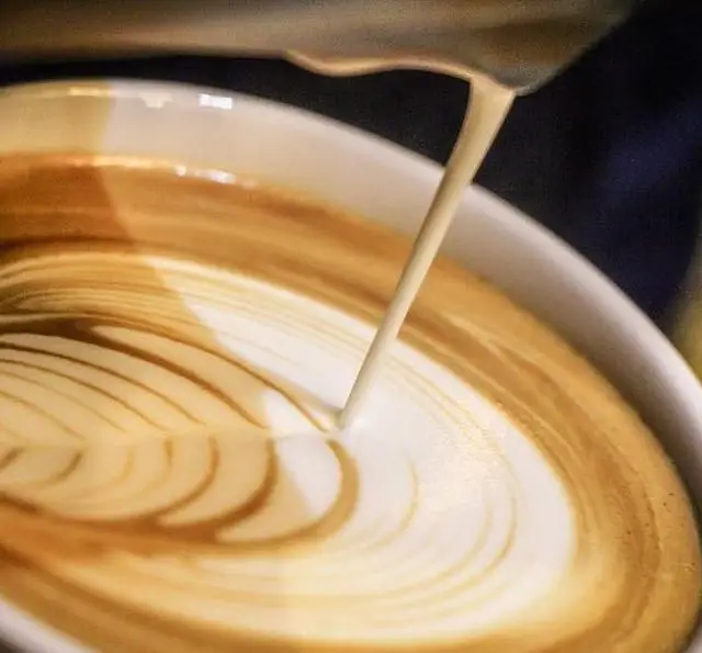 تصویری جذاب از ریزش شیر و فرم‌دهی لاته آرت گل در فنجان قهوه در کافه رستوران داریما، نمادی از مهارت باریستا و کیفیت بالای نوشیدنی.