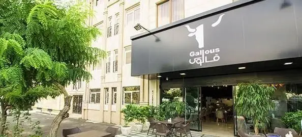 نمای بیرونی رستوران فرنگی قالوس، با تابلوی سیاه رنگ، لوگوی گاو و فضای سبز دلنشین. میز و صندلی در فضای باز آماده پذیرایی.