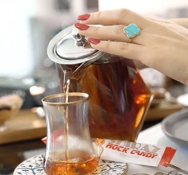 ریختن چای گرم از قوری شیشه‌ای به استکان سنتی توسط دستی با انگشتر فیروزه، در محیطی دلنشین از کافه رستوران عربی مدائن، با نبات شیراز.