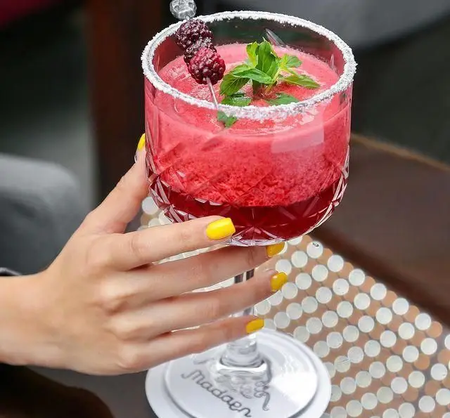نوشیدنی کوکتل میوه ای خنک و قرمز در لیوان طرح دار با لبه شکر، تزئین شده با توت و نعنا در کافه رستوران عربی مدائن.