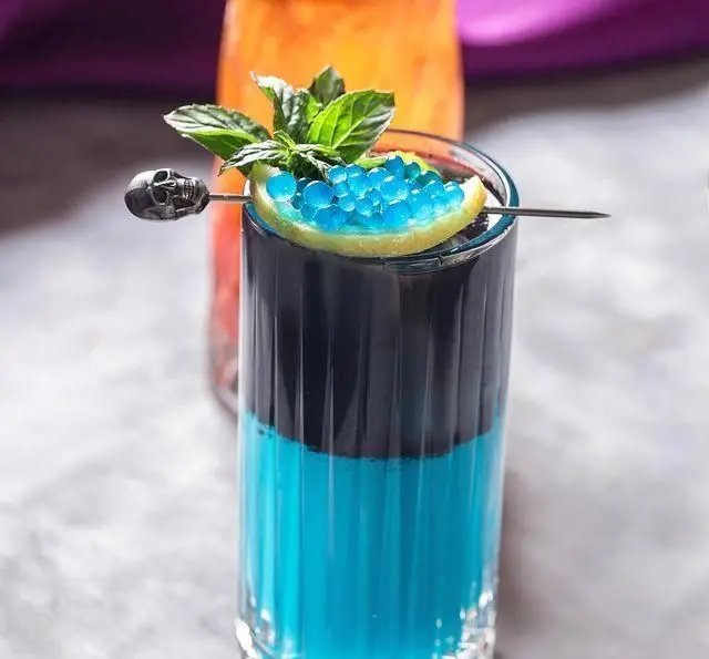 نوشیدنی فانتزی و خاص کافه رستوران عربی مدائن با لایه‌های آبی و مشکی، تزیین شده با بابلس آبی و اسکلت برای تجربه‌ای متفاوت.